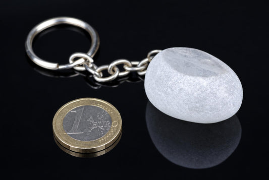 A silver-toned keychain with a smooth, pale grey stone pendant and chain rests on a dark reflective surface next to a one-euro coin.