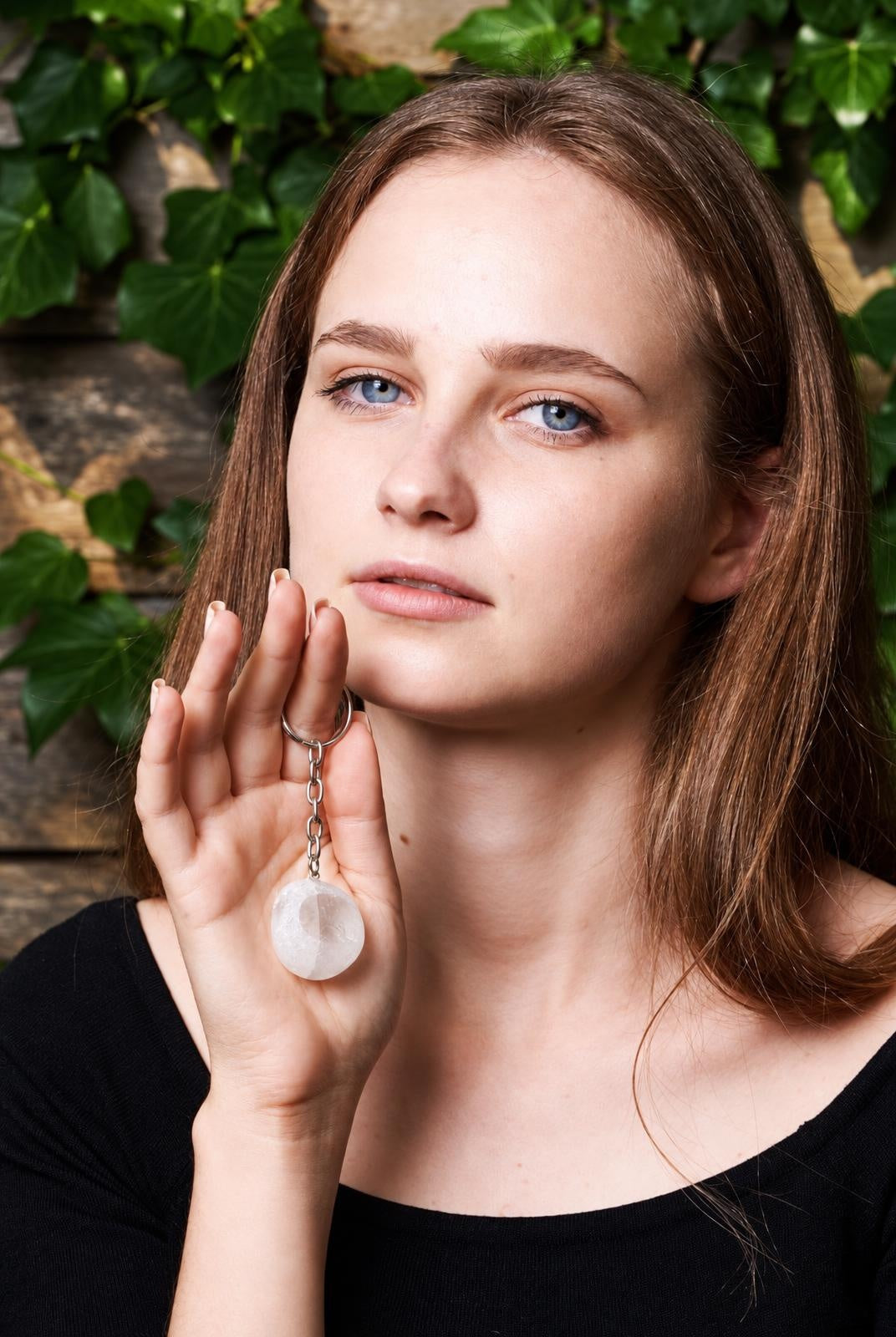 A woman holds a silver-toned keychain with a round, translucent white quartz pendant against a dark surface and green leaves.
