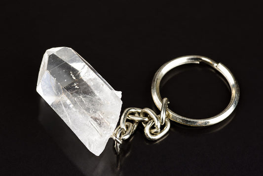 A silver-toned keyring with a chain connects to a clear, faceted quartz crystal on a dark surface.