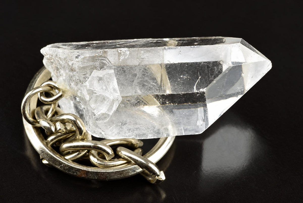 A clear quartz crystal point attached to a silver-toned metal keyring, resting on a dark surface.