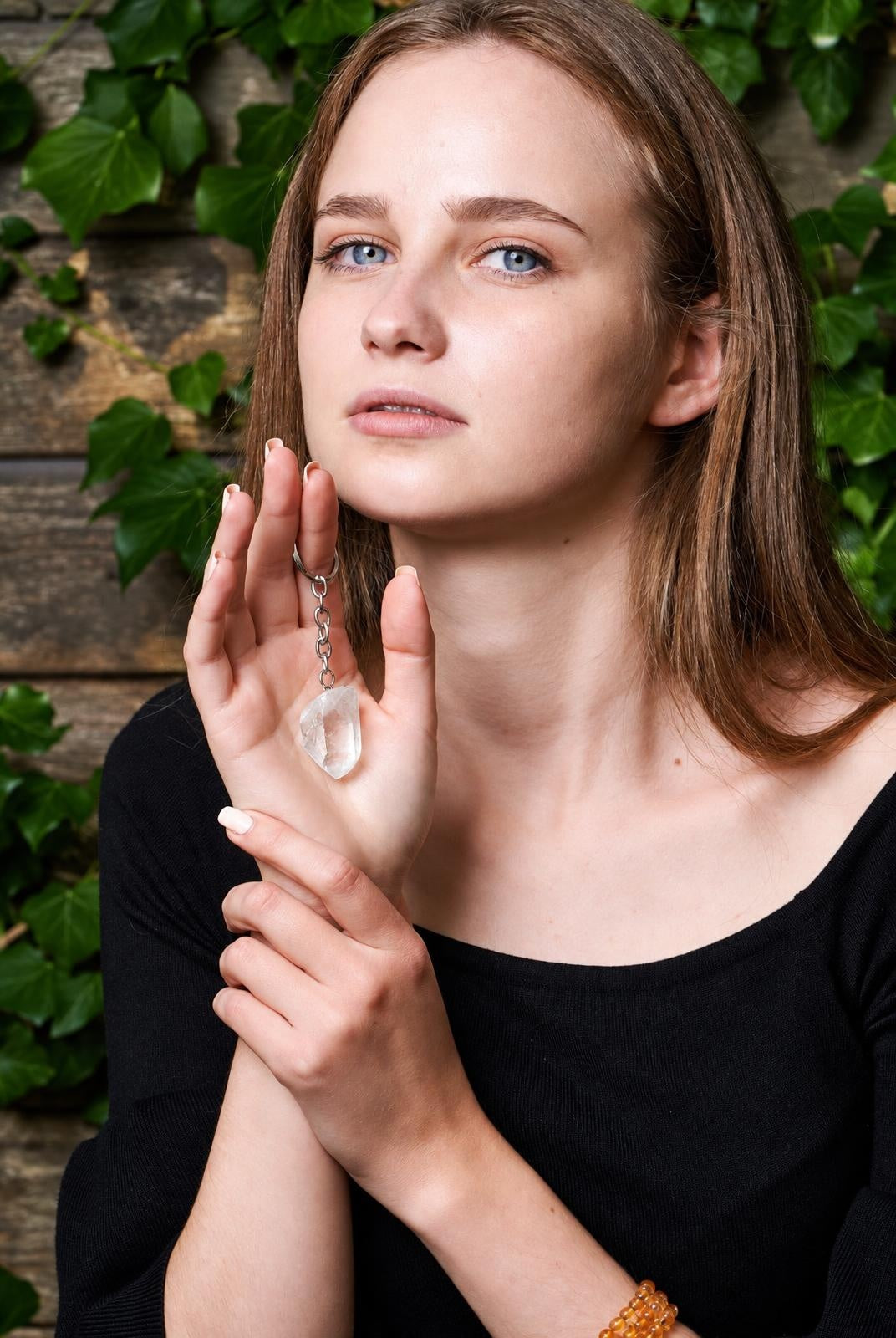 A woman holds a silver-toned keyring with a clear quartz crystal pendant against a wooden surface and green ivy.