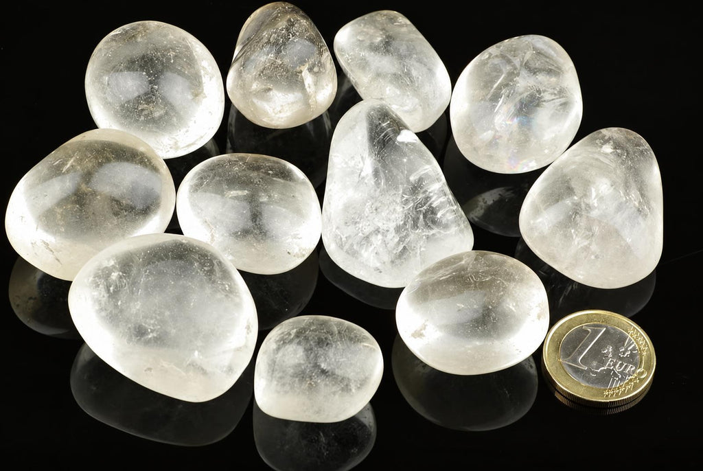A group of smooth, clear quartz stones on a dark surface with a one-euro coin for scale.