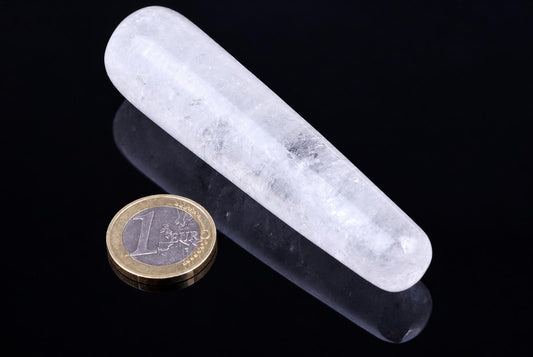 A translucent white quartz massage stick lies diagonally on a dark, reflective surface next to a one-euro coin.