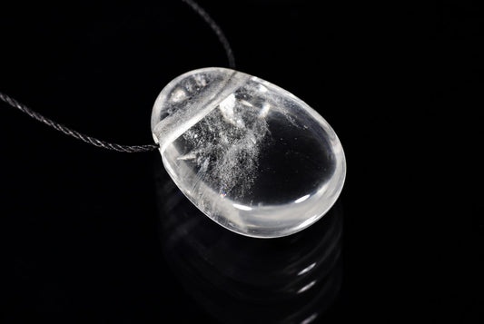 A clear quartz pendant with internal white inclusions hangs on a dark cord against a black reflective surface.