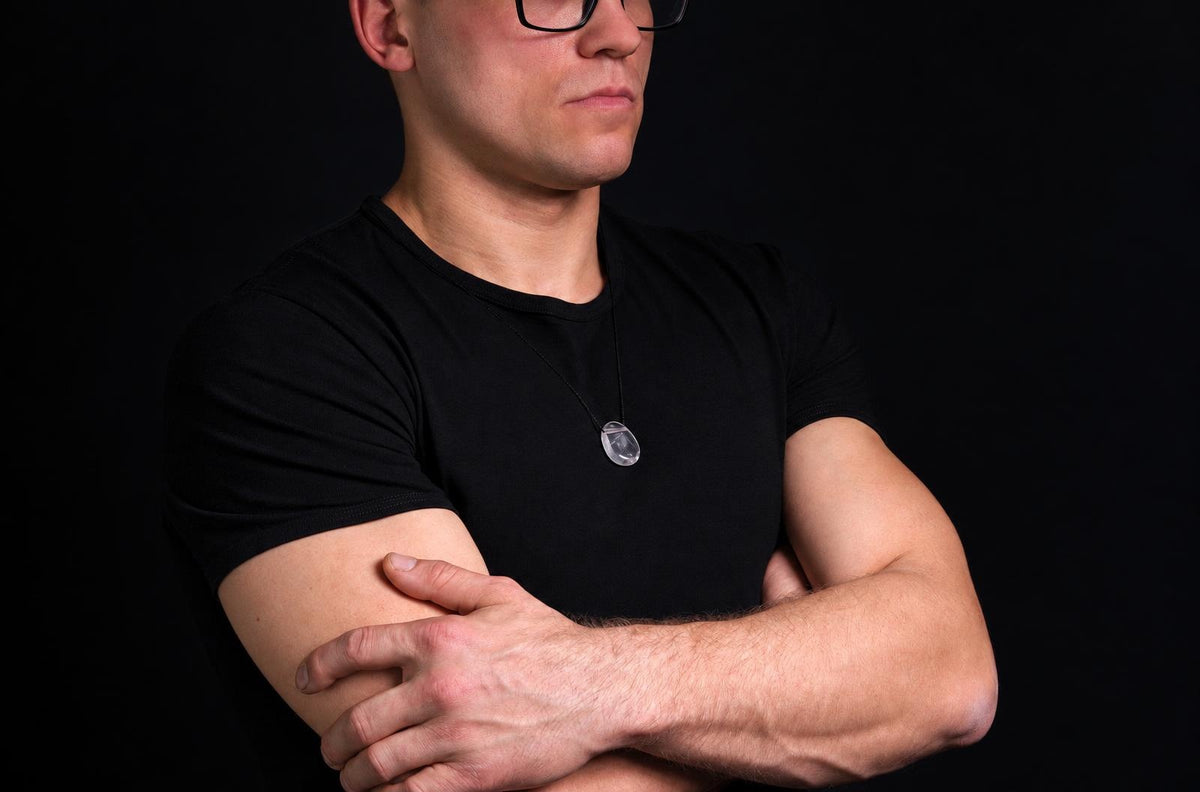 A man in a black t-shirt wears a necklace with a clear, faceted quartz pendant on a dark surface.