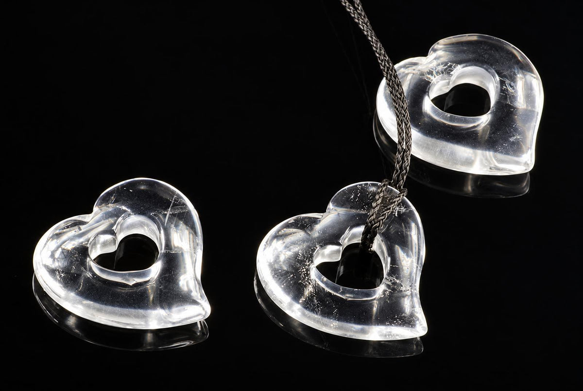 Three clear, heart-shaped quartz pendants with central holes rest on a dark surface; one hangs from a braided cord.