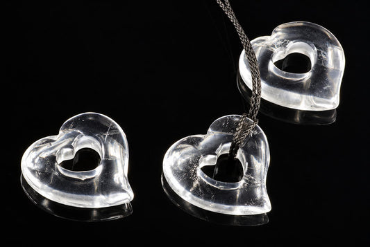 Three clear, heart-shaped quartz pendants with central holes rest on a dark surface; one hangs from a braided cord.