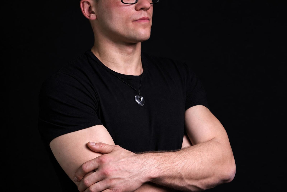 A man in a black t-shirt wears a silver-toned heart-shaped pendant on a dark surface.