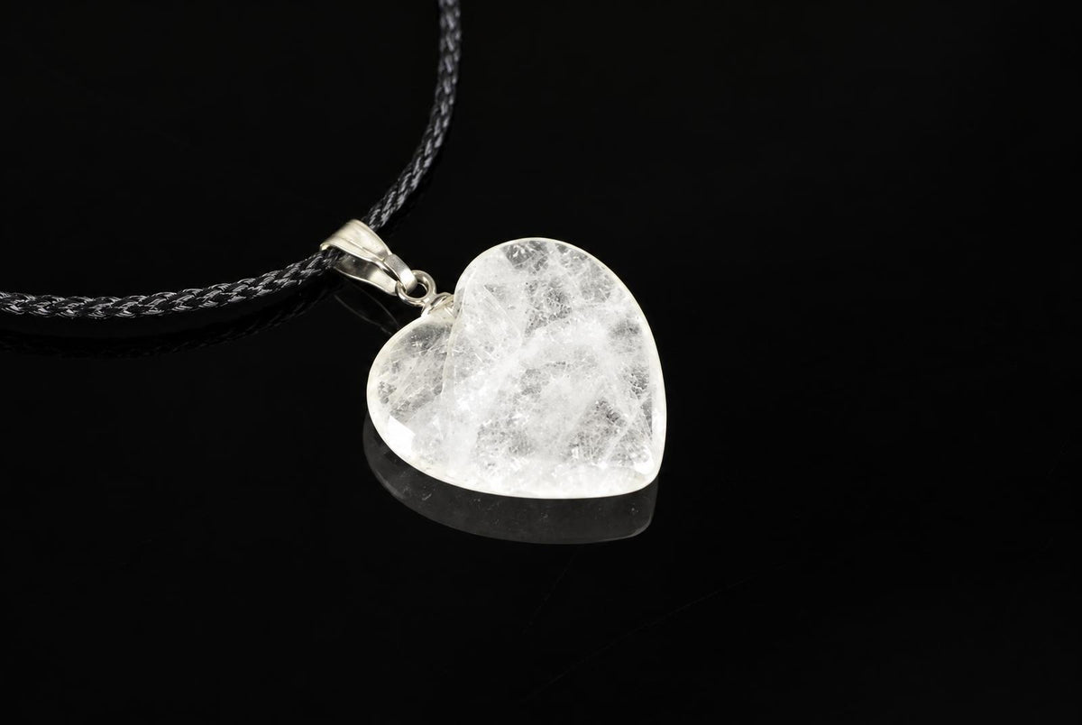 A heart-shaped quartz pendant with a silver-toned bail hangs from a dark braided cord against a black reflective surface.