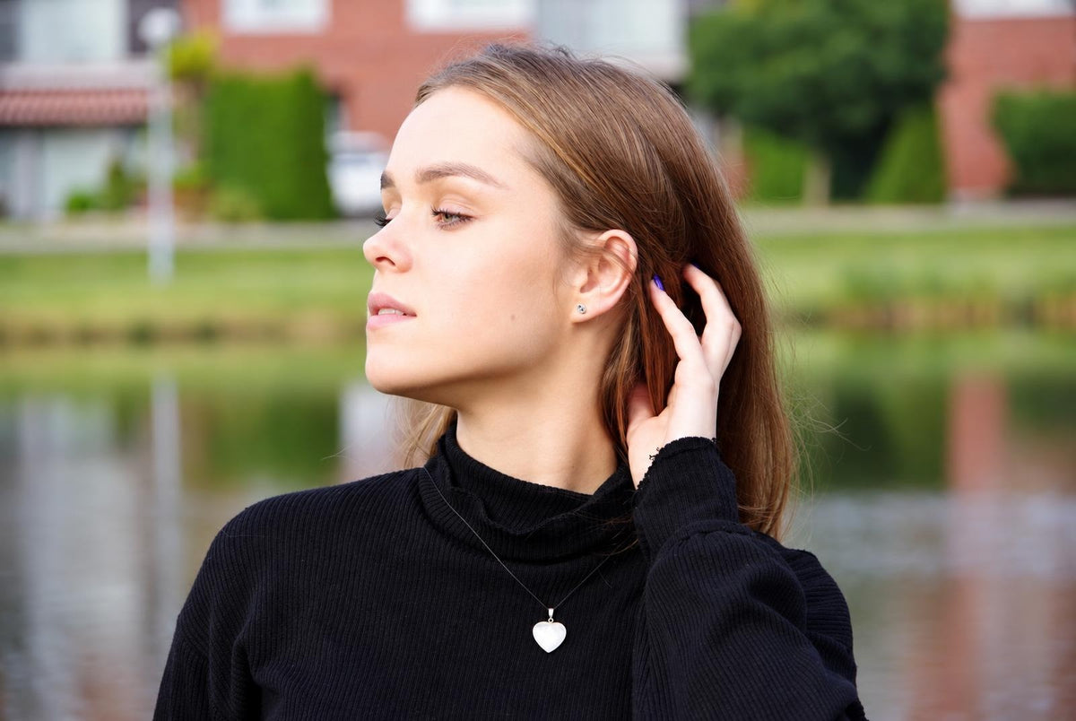 A woman wears a black turtleneck and a silver-toned heart-shaped pendant necklace against a blurred outdoor background.