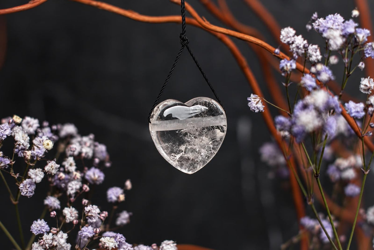 A clear, heart-shaped quartz pendant hangs from a black cord against a dark background with purple flowers and brown branches.