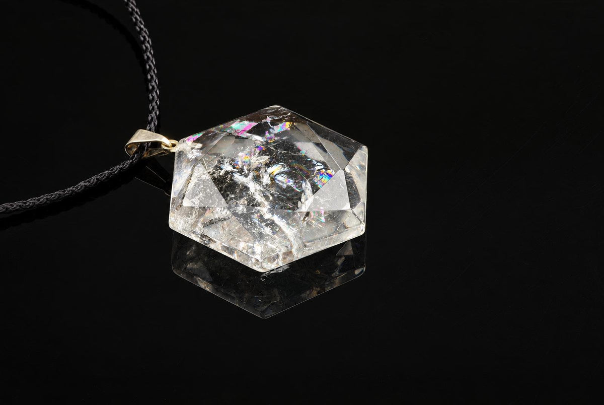 A clear quartz pendant has a faceted hexagonal shape and visible internal markings, hanging from a black cord above a dark reflective surface.