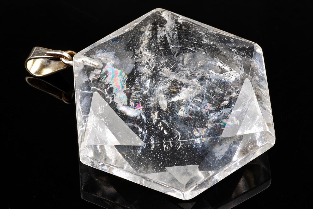 A clear, hexagonal quartz pendant with internal inclusions and a gold-coloured bail rests on a dark surface.