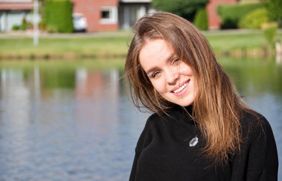 A smiling woman with brown hair wears a black top and a clear hexagonal quartz pendant necklace, standing outdoors near water.