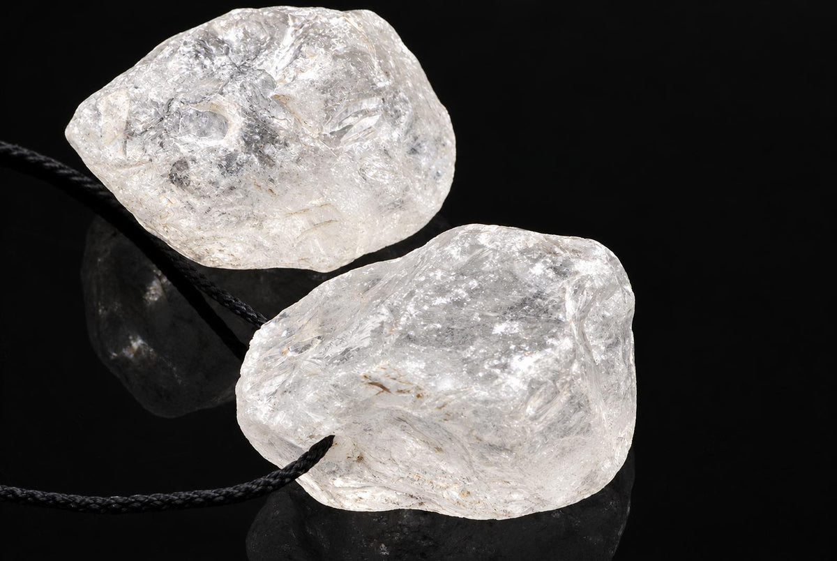 Two clear quartz stones on black cords, resting on a dark surface against a black background.