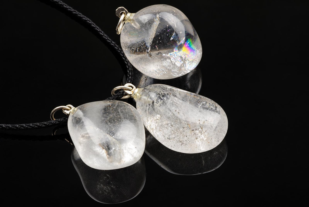 Three clear quartz pebble pendants with gold-toned fittings on black cords, resting on a dark reflective surface.