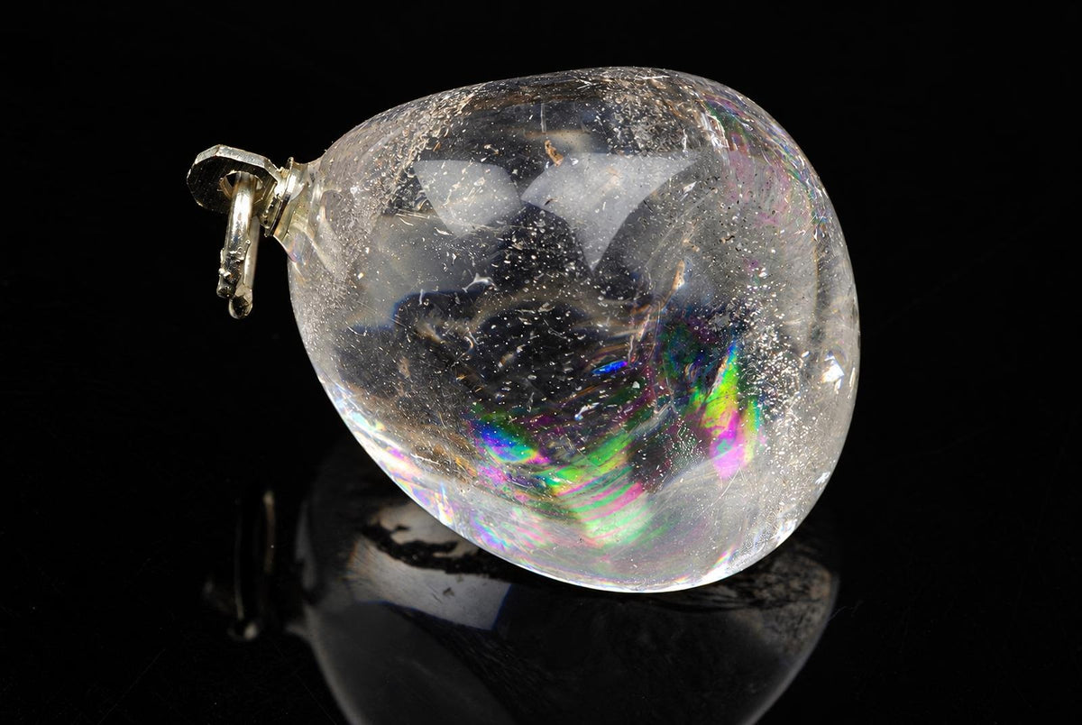 A clear quartz pendant with a silver-toned bail, showing internal inclusions and rainbow iridescence on a dark surface.