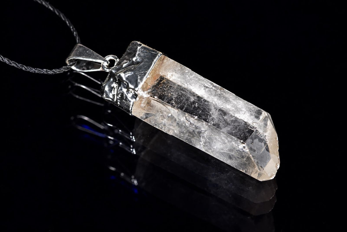 A clear pointed quartz pendant with visible internal textures and a decorative silver-colored cap hangs from a black cord against a dark background.