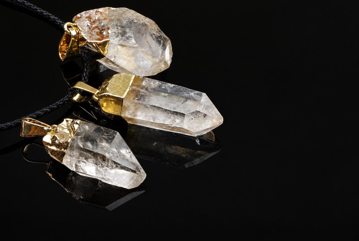 Three clear quartz crystal pendants with gold-toned settings on dark cords, arranged diagonally on a reflective black surface.
