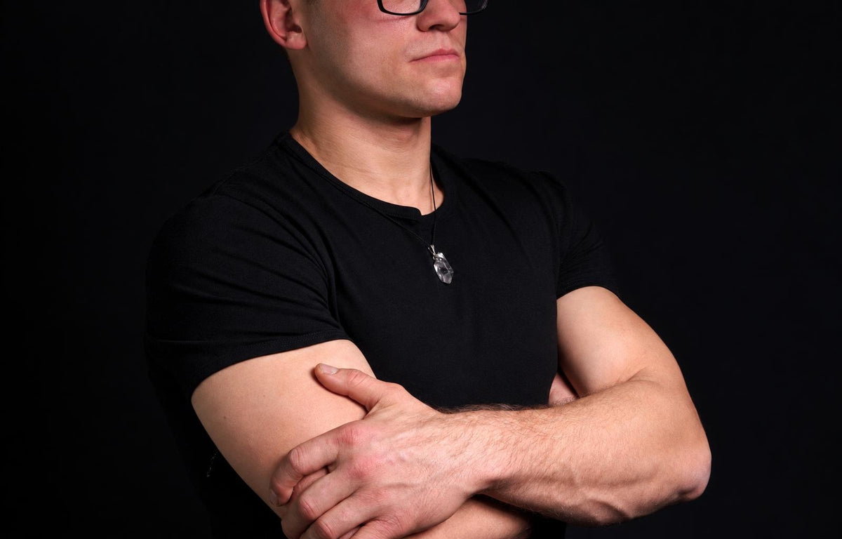 Man in black t-shirt with crossed arms wears a clear quartz pendant on a dark surface.