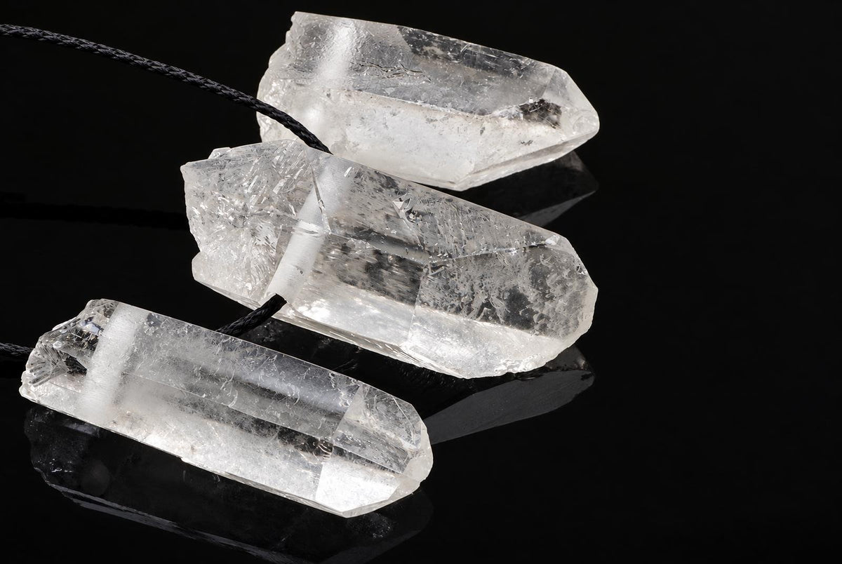 Three clear quartz crystal pendants with black cords rest on a dark, reflective surface.