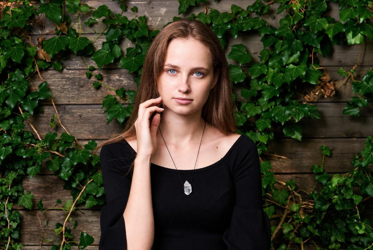 A woman wears a black top and a necklace with a clear quartz pendant on a dark wooden surface covered in green ivy.