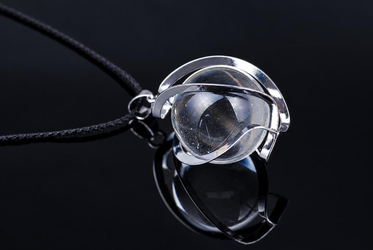 A silver-toned pendant with a clear quartz sphere, wrapped in metal bands, hangs from a black cord against a dark surface.