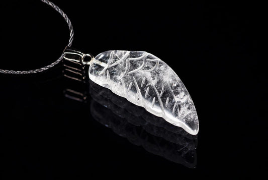 A clear quartz pendant shaped like a wing with white inclusions, attached to a silver-toned cord on a dark reflective surface.