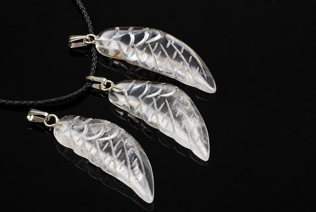Clear quartz wing pendants with carved feather grooves and silver-tone loops are arranged with black cords on a glossy dark surface.