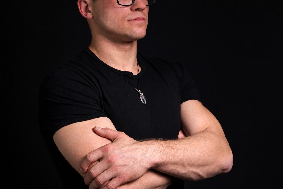 A man in a black t-shirt wears a silver-toned pendant with a clear quartz crystal on a dark surface.