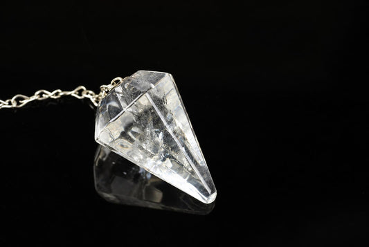 A clear quartz pendulum with a pointed, faceted shape hangs from a silver-toned chain on a dark surface.
