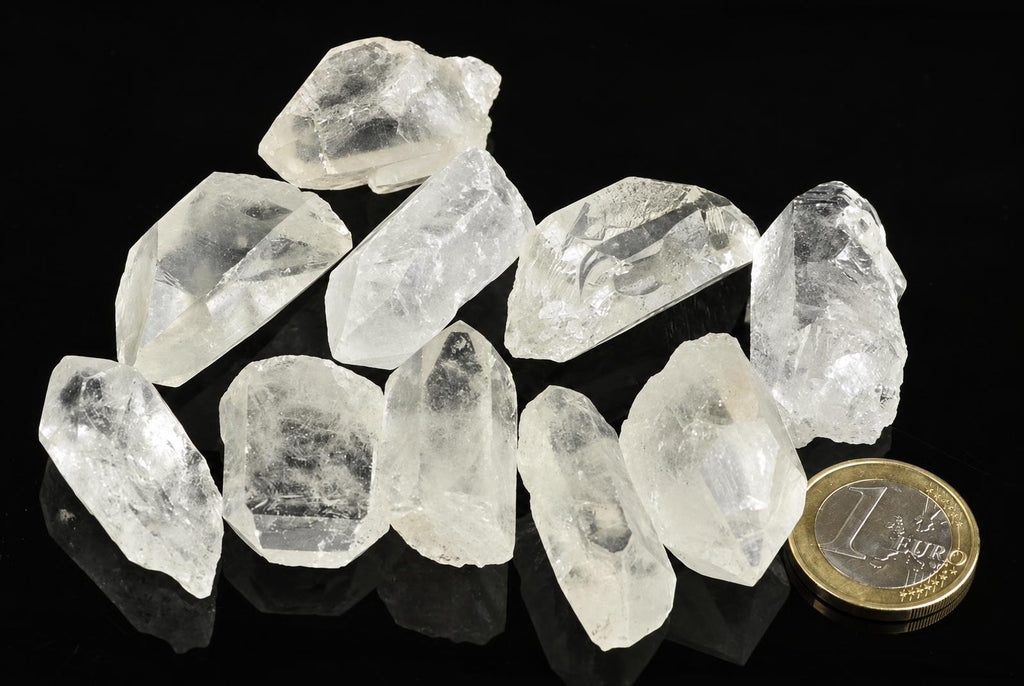A group of clear, translucent quartz crystals with pointed tips arranged on a dark surface next to a one-euro coin.