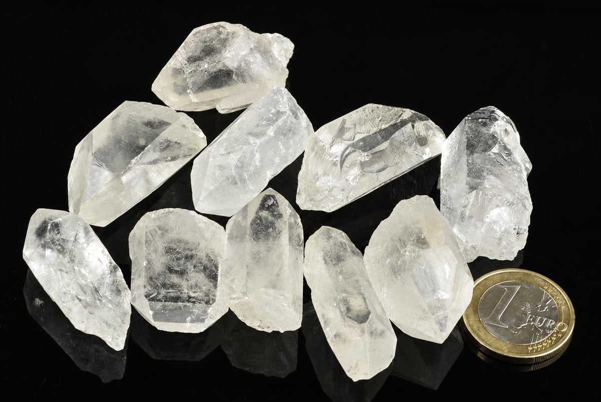 A group of clear, translucent quartz crystals with pointed tips arranged on a dark surface next to a one-euro coin.