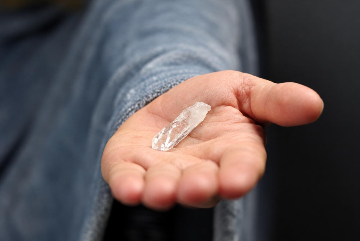 A person holds a small, clear quartz crystal point in their open palm against a dark background.
