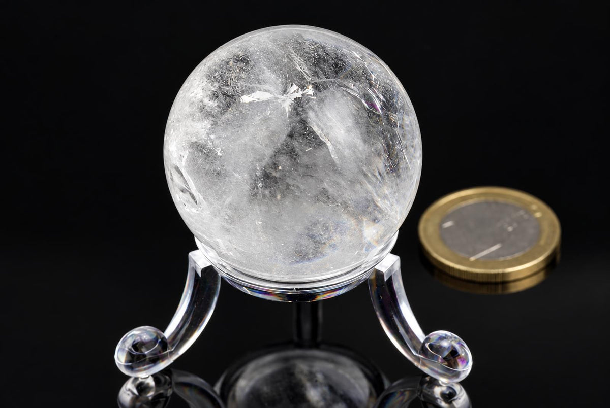 A clear quartz sphere with internal inclusions sits on a transparent stand against a dark surface, next to a coin.
