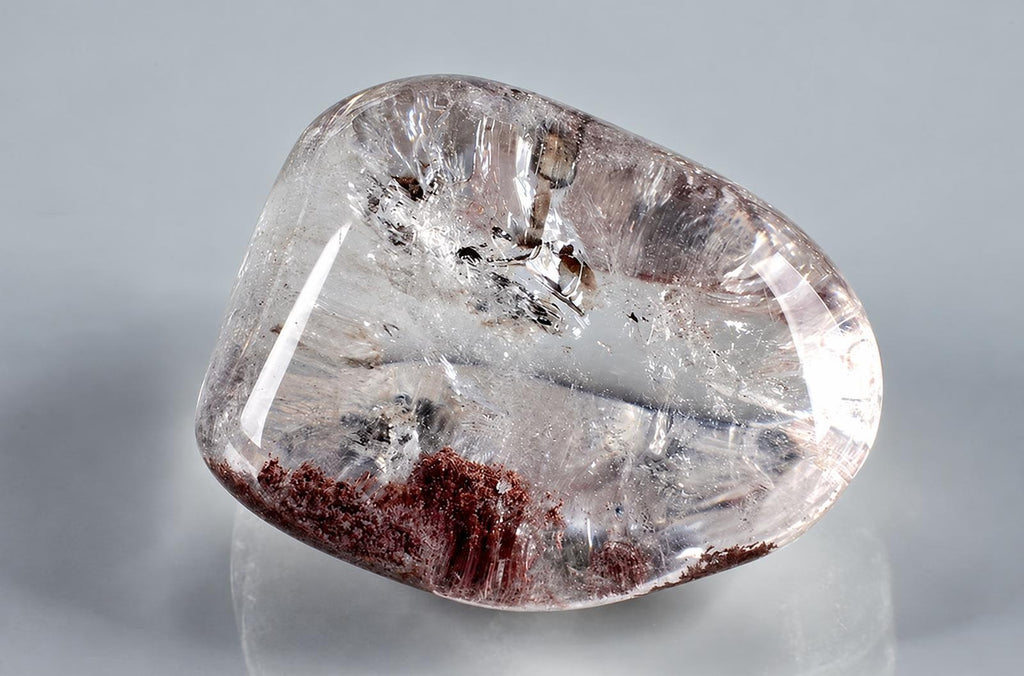 A polished, transparent quartz stone with dark brown inclusions at the base and internal crystalline structures on a pale surface.