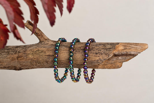 Three rainbow-colored beaded bracelets rest on a rough wooden branch against a pale background with red leaves above.