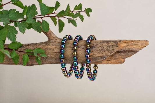 Three rainbow-colored beaded bracelets rest on a rough wooden branch with green leaves against a pale background.
