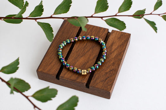 A bracelet of shiny multicolored round beads with a small square metal bead rests on wooden slats beneath a leafy branch.