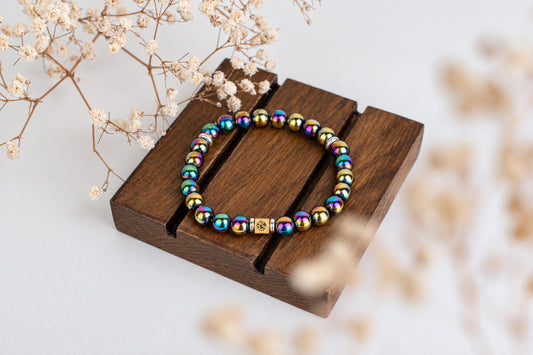 A bracelet with rainbow-colored spherical beads and a gold-toned square charm rests on a dark wooden surface.