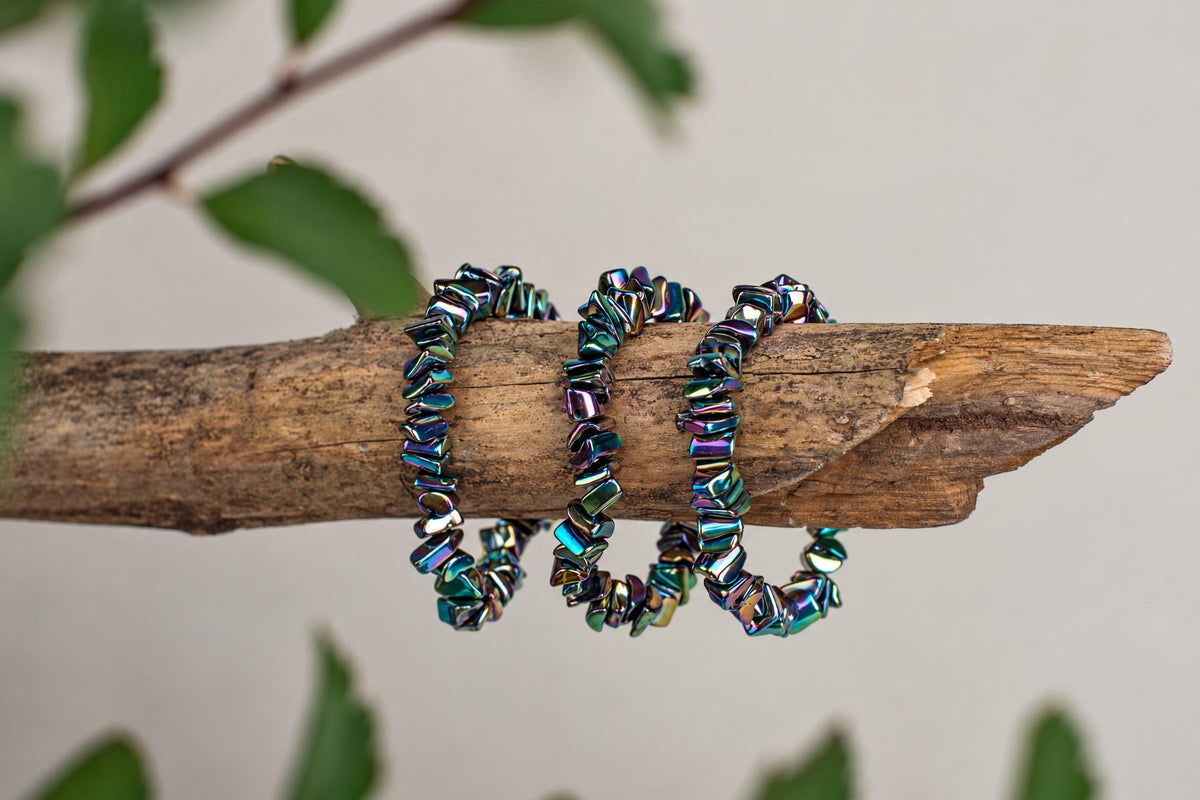 Three rainbow-hued chip bracelets rest on a rough wooden branch, with blurred green leaves in the background.