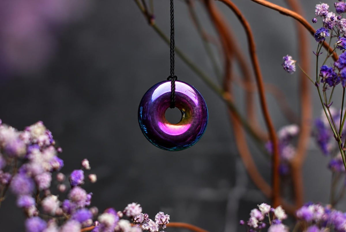 A donut-shaped pendant with a rainbow iridescent finish hangs from a black cord against a dark background with purple flowers.