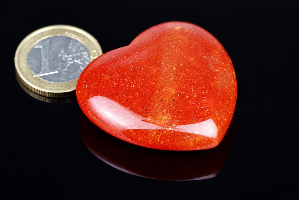 A polished red heart-shaped stone with a speckled texture sits on a dark surface next to a one-euro coin.