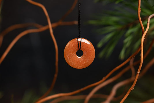 A round, orange-brown pendant with a central hole hangs from a black cord against a dark background with blurred branches.
