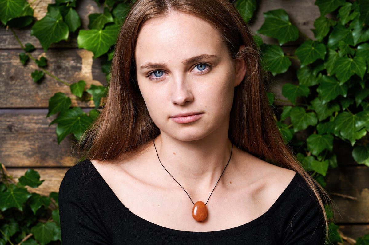 A woman wears a black top and a necklace with an oval, orange-brown pendant on a thin black cord against a wooden wall with green ivy.