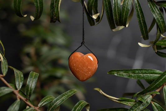 A polished, heart-shaped red aventurine pendant hangs from a black cord against dark green leaves.