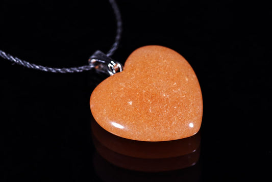A polished heart-shaped pendant in orange-brown aventurine with a silver-toned bail and dark cord, resting on a black reflective surface.