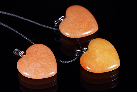 Three heart-shaped pendants in orange and yellow tones on dark cords, resting on a reflective black surface.