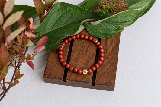 A red beaded bracelet with a square gold-toned charm rests on a dark wooden surface, surrounded by green and reddish leaves.