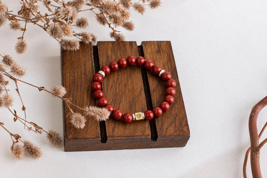 A bracelet with red round beads and silver-toned accents rests on a dark wooden surface, surrounded by dried flowers.